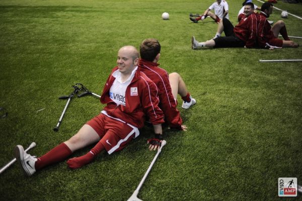 Na zdjęciu piłkarze siedzący na boisku plecami do siebie rozciągają się.