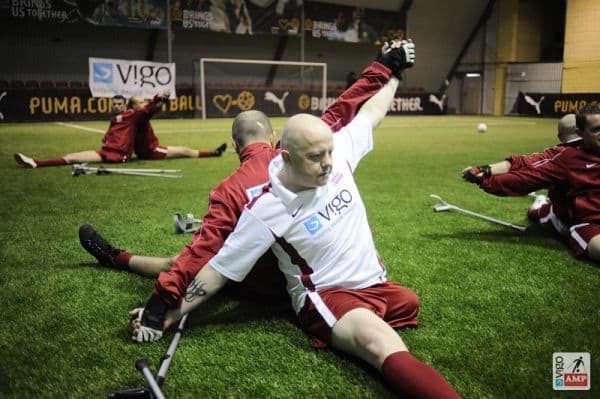 Na zdjęciu Maciej Kosiorek w białej bluzie sportowej rozciąga się z kolegą. Oboje siedzą na boisku plecami do siebie. Chwycili się za wyprostowane w górę ręce i wzajemnie się rozciągają.