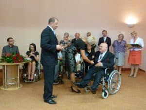 Na zdjęciu Andrzej Szymczakowski na wózku wśród ludzi odbiera nagrodę z rąk mężczyzny stojącego przed nim.