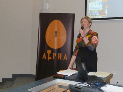 Na zdjęciu Anna Prokopiak w krótkich blond włosach mówi do mikrofonu podczas wystapienia. W tle za nią prezentacja multimedialna, na lewej stronie zdjęcia roll-up Alpha
