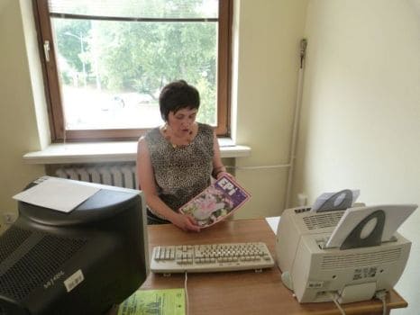 Na zdjęciu Barbara Lisek za biurkiem na którym stoi sprzęt komputerowy - klawiatura, duży monitor i drukarka. Prezentuje do zdjęcia jaki obraz plastyczny, w tle duże okno.