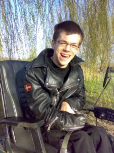 Portet Daniela Szarpaka. Chłopak w krótkich włosach, w okularach z szerokim uśmiechem w czarnej skórzane kurtce siedzi na wózku inwalidzkim. W tle wierzba