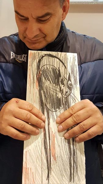 Na zdjęciu Jarosław prezentuje dzieło namalowane na wąskim kawałku papieru pt. Kot - prezentuje ono czarnego kota o długich łapach z ogonem wykręconym do góry kierującym się łukowano ku łebkowi kota.