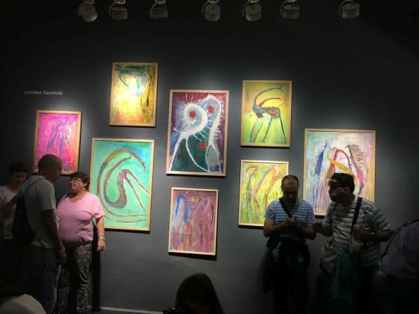 Na zdjęciu na czarnej, oświetlonej reflektorami ścianie galerii wiszą obrazy autorstwa Jarosława. Wernisaż wystawy pod tytułem "Dużo pracy" Obrazy przedstawiają różno-kolorowe interpretacje żyrafy przez autora