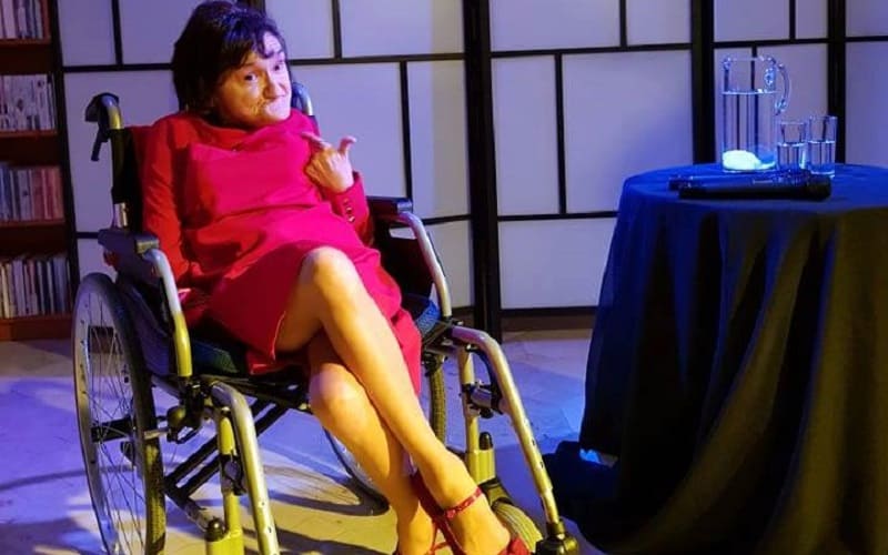 Na zdjęciu Joanna Pąk w różowym kostiumie - sukience siedzi na wózku inwalidzkim, w blasku reflektora.