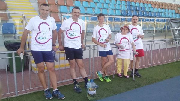 Na zdjęciu Krystyna w żółtych spodniach stoi z grupą młodych ludzi na tle trybun. Grupa opiera się o barierki, wszyscy mają takie same białe koszulki z logiem maratonu w kształcie serca.