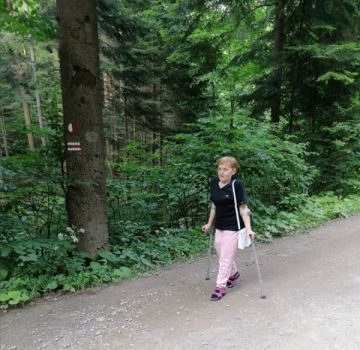 Na zdjęciu Krystyna idzie szlakiem w Beskidzie Niskim o kulach na tle gęstego zielonego lasu.
