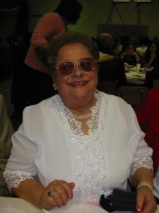 Portet zdjęciowy Teresy Ciodyk. Starsza kobieta w krótkich siwyhc włosach w przyciemnionych okularach i w białej bluzce w koronkę uśmiecha się do fotografa