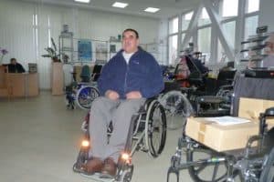 Na zdjęciu Tadeusz Ćwikła siedzący na wózku inwalidzkim. Czarne włosy na jeża, ookulary, granatowy polar, szare spodnie i brązowe skórzane buty. Siedzi wśród wózków w sklepie rehabilitacyjnym.