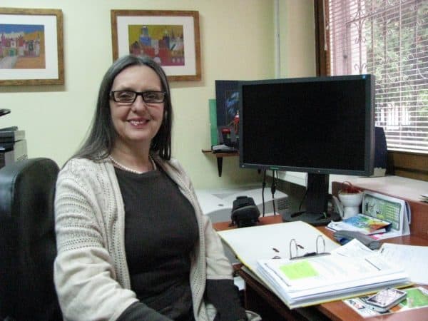 Na zdjęciu Zofia Brzozowska w czarnych okularach uśmiecha się do zdjęcia siedząc przy biurku z komputerem i dokumentami.