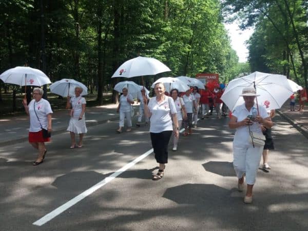 Na zdjęciu Amazonki podczas marszu przez park. Ubrane na baiało trzymają w dłoni otwarte białe parasolki.