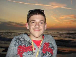 Portret zdjęcuiowy Wojtka Mazura. Umiechnięty chłopak z lekkim wąsem w szarej bluzie i w okularach na głowie. Siedzi na tle morza przy zachodzącym słońcu.