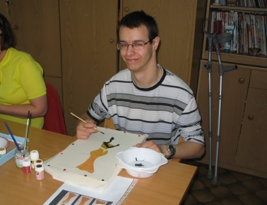 Kamil w okularach podczas malowania pracy przy stole. Uśmiecha się do zdjęcia