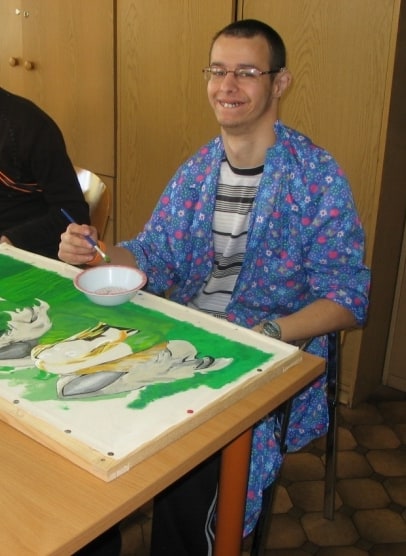 Kamil podczas malowania, siedzi w niebiskim fartuchu z pędzlem w ręku