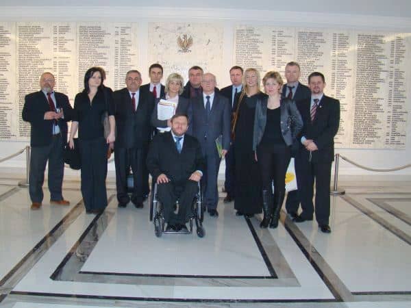 Grupa ludzi w tym mężczyzna na wózku w eleganckich strojach pozuje na tle pamiątkowej tablicy w Sejmie RP.