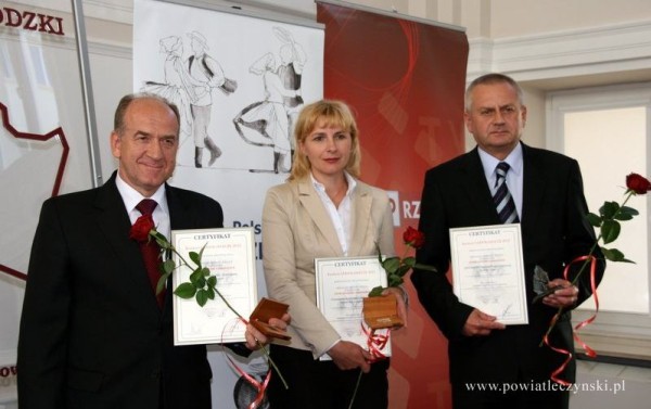 Małgorzata Paprota na środku pozuje z dwoma mężczyznami po obu jej stronach. W dłoniach trzymają laury, dyplomy uznania i różę.