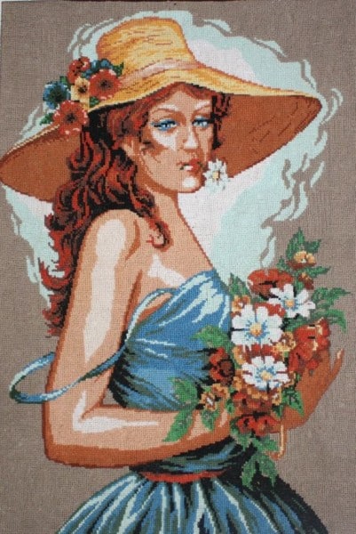 Obraz haftowany. Kobieta w kapeluszu słomkowym w niebieskiej sukience z bukietem kwiatów w dłoni.