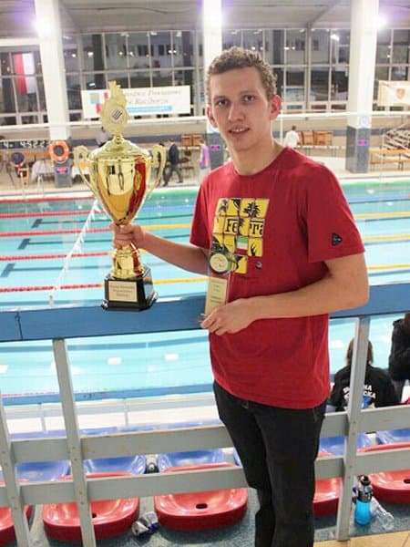 Artur piórko pozuje z trybun z pucharem na tle basenu pływackiego.