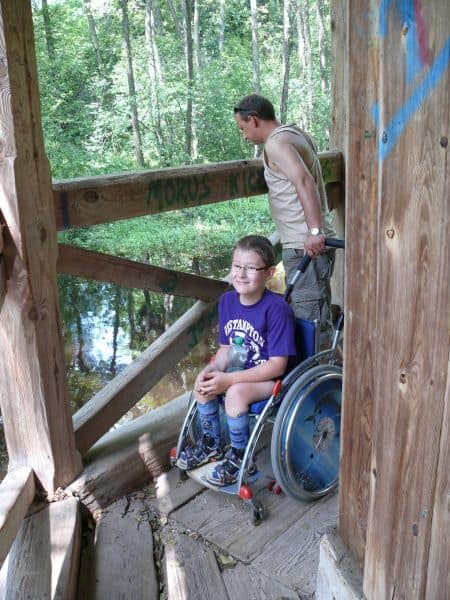 Dziecko na wózku na tarasie okalającym kapliczkę nad rzeką. Przez barierkę wygląda mężczyzna