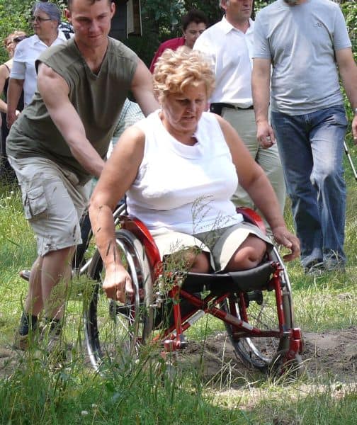 Kobieta na wózku jedzie przez zapiaszczoną ścieżkę. Pomaga jej młody chłopak.