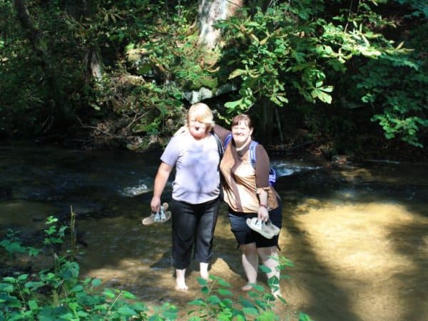 Dorota Szpak z mamą pozują do zdjecia stojąc po kostki w wodzie rzeki płynącej w lesie.
