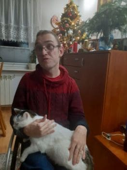 Michał Wożniak z białym kotem na kolanach, na wózku. UJbrany w czerwoną bluzę. Nosi okulary