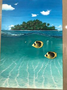 Obraz. Dolna połowa obrazu pod wodą, widać piach i blask fal na dnie, pływają dwie ryby. Góna połowa widok na małą wysepkę pełną palm na tle błękitnego nieba,