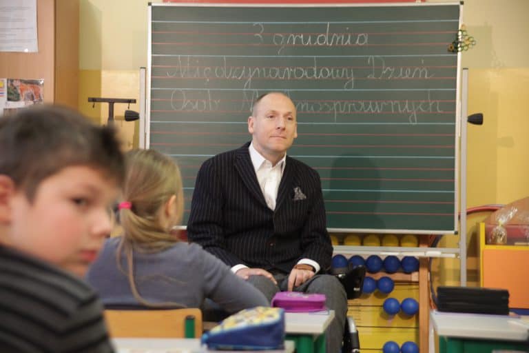 Piotr Pawłowski prowadzi w szkole zajęcia dla dzieci. W tle tablica z napisem: 3 grudnia Międzynarodowy Dzień Osób Niepełnosprawnych