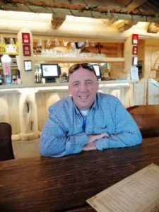 Piotr Gruszecki w jeansowej koszuli, w okularach przeciwsłonecznych na głowie uśmiecha się do zdjęcia siedząc w restauracji.