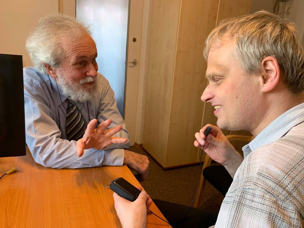 Dwóch mężczyzn rozmawia. Są uśmiechnięci. Oboje są wyposażeni w aparaty słuchowe i pętle indukcyjne.