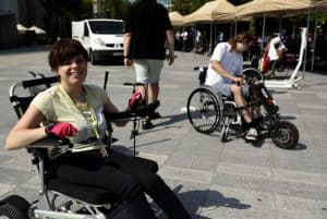 kobieta siedzi na wózku inwalidzkim. Uśmiecha się do zdjęcia. W tle mężczyzna na wózku. Są na deptaku w mieście.