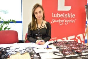 kobieta siedzi przy stole z ulotkami i brzoszurami. W tle za nią baner reklamowy stojący.