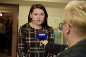 Kobieta stoi i ma przed sobą mikrofon z napisem TVP3 Lublin. Jest nachylona do mikrofonu, który trzyma kobieta. Kobieta z mikrofonem stoi tyłem do obiektywu i nie widać pełnej sylwetki.