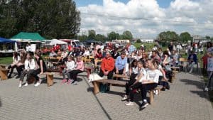 Grupa osób siedzi na drewnianych ławkach na świeżym powietrzu. Po lewej stronie na trawniku stoi kilka namiotów.