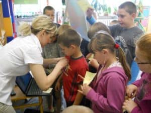 Kobieta przypina przypinkę chłopczyjowi. Dzieci wokół czekają na swoja kolej