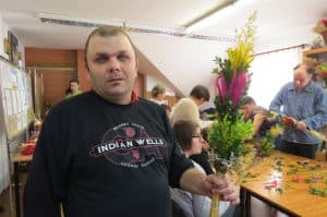 Mężczyzna stoi na wprost do zdjęcia. W dłoni trzyma kolorową palmę. W tle stół z materiałami do robienia palemek oraz inni ludzie.