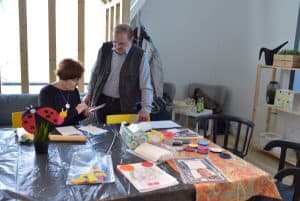 Kobieta siedzi na krześle przy dużym stole, na którym są farby, książka. Trzyma telefon w rękach a mężczyzna stoi koło niej i patrzy na nią.