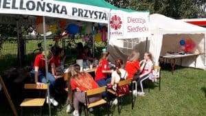 Na krzesłach siedzi kilka osób- dziewczynki i wolontariusze. Po prawej stronie dziewczynka siedzi na krześle a kobieta kuca przed nią i dotyka jej twarzy. Nad nimi jest napis “Caritas diecezja siedlecka”