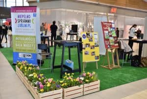 Plakat informacyjny: niepełnosprawnilublin.pl. Wokół niego są kwiaty w skrzynkach i sztuczna trawa na której stoją inne plakaty.