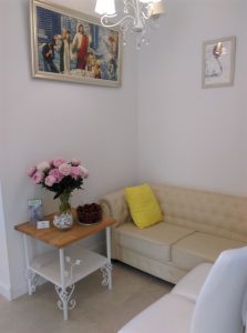 Biały pokój urządzony w pastelowych kolorach. Po lewej stronie jest stoliczek na którym stoi wazon z różowymi piwoniami. Obok stoi biała kanapa, a na ścianach wiszą obrazy.