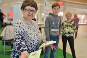 Kobieta trzyma w rękach ulotki, a w jedną z nich wyciąga w kierunku obiektywu. Koło niej stoi młody mężczyzna i kobieta.