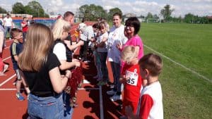 Grupa osób stoi na bieżni. Niektórzy mają na koszulkach przyklejone numerki. KObiety po lewej stronie niosą w rękach pęk medali.