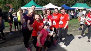 Grupa osób pozuje do zdjęcia. Część osób pokazuje swoje bicepsy. Wszyscy szeroko się uśmiechają, a za nimi są inne osoby i namioty.