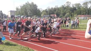 Na bieżni jest duża grupa osób. Wszystkie osoby są w pozie poruszania się. Biegną kobiety, mężczyźni, dzieci.