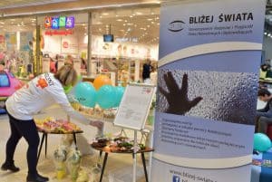 Baner z napisem: “Bliżej świata”. Po lewej stronie dwa małe stoliki nad którymi pochyla się kobieta. Przed nią są niebieskie balony.