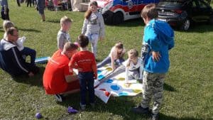 Na trawie dzieci grają w grę Twister. Wokół nich są dorośli i dzieci, którzy obserwują. Za nimi stoją samochody i karetka, które są ucięte.