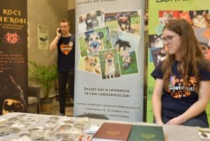 Po prawej stronie stoi kobieta. Przed nią jest stół a na nim leżą książki. Za kobietą są bilbordy, a po lewej stronie jest mężczyzna, który rozmawia przez telefon.