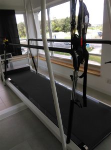 Długa bieżnia z poręczami stojąca przy oknie. Na jej końcu wysoki ekran.