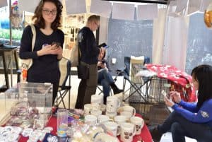 Kobieta stoi i patrzy na stół na którym są różne kubki. Po prawej stronie siedzi kobieta. Na stole po lewej stronie jest skrzynka w której są pieniądze.