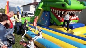 Dzieci są na dmuchanej zjeżdżalni, na której jest głowa smoka. Po lewej stronie stoją dorośli. Na trawie przed zjeżdżalnią siedzi mała dziewczynka.